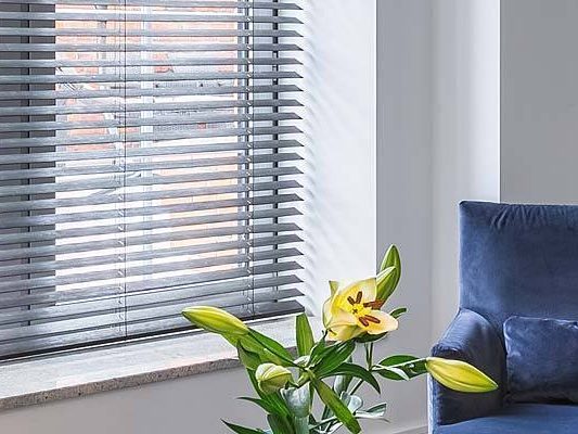a picture of Lily flowers in a vase beside a blue chair by a window with venetian blinds.